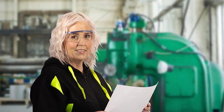 En blond kvinna med skyddsglasögon och iklädd varselkläder står i en industri