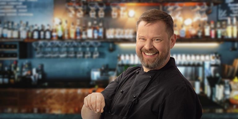 En man med skägg, iklädd svart kockskjorta står bakom en bar